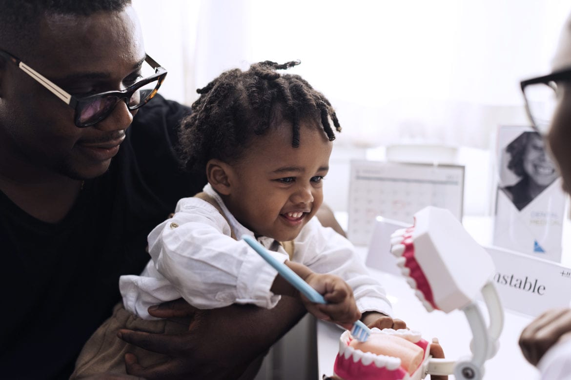 Son being held by his father while he is playing with toothbrush and model set of teeth