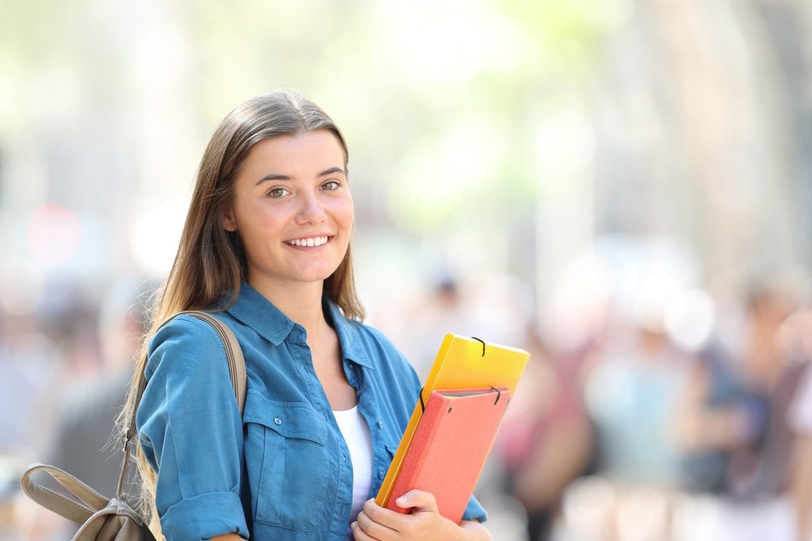 A female college student is holding a notebook and bookbag.