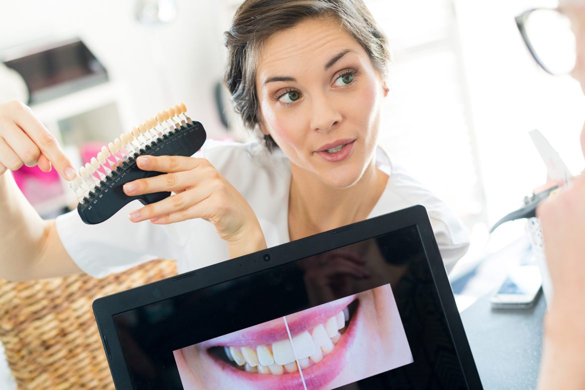 blog-eds-cosmed-dent-cost Dentist holding tooth samples for bleaching treatment while patient looks at a smile within a picture frame
