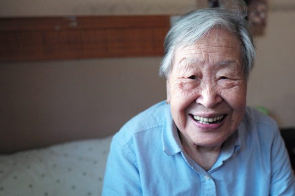 An elderly woman smiles while sitting on furniture.
