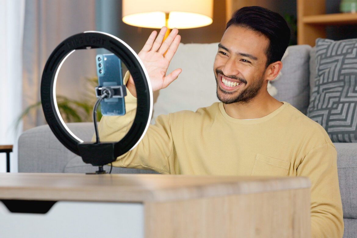 A man is smiling in front of his phone and ring light.