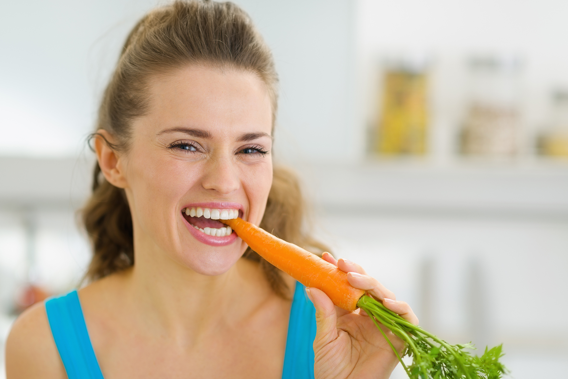 A woman is smiling while eating a carrot.